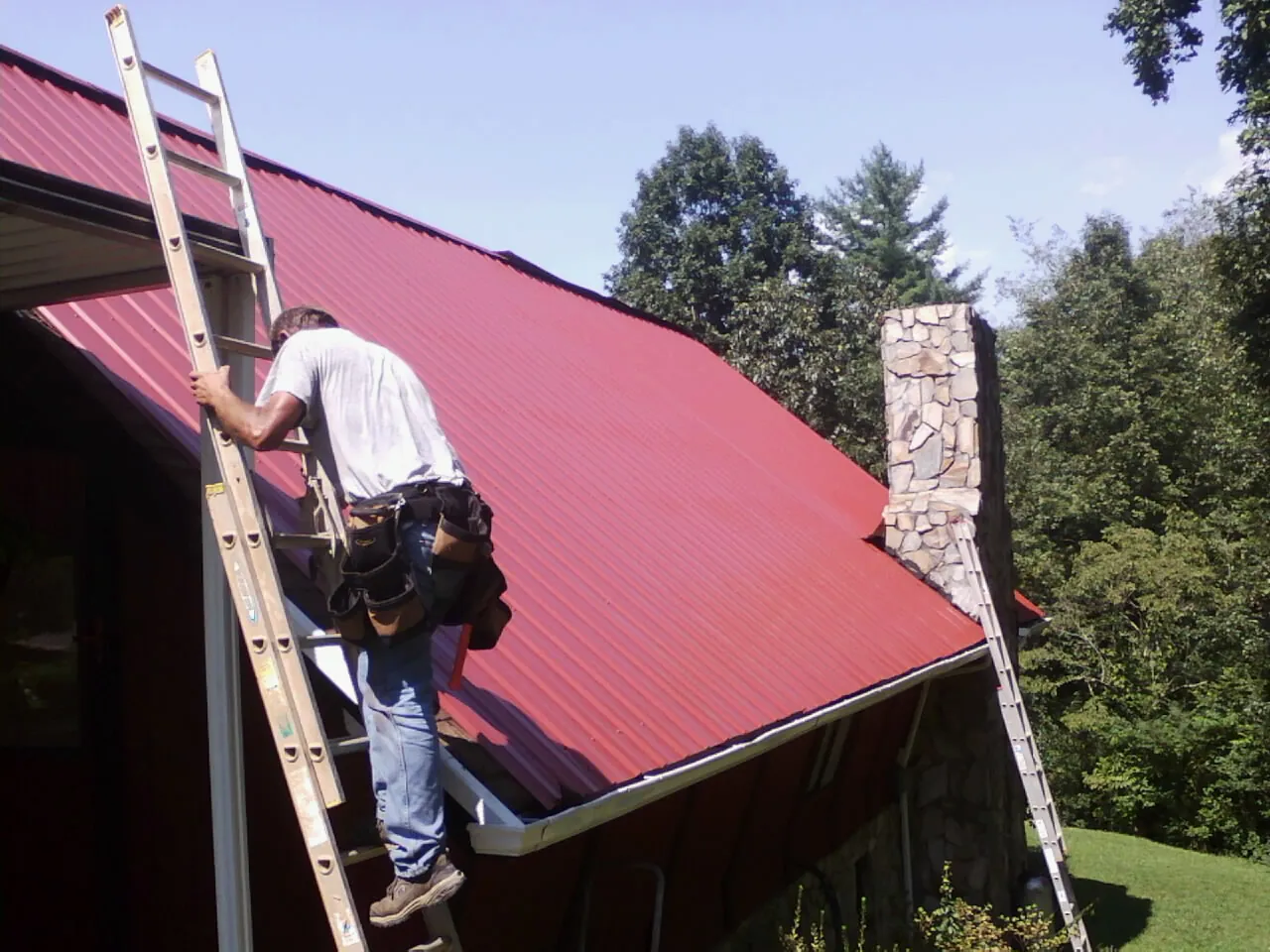 Roofer on a ladder beside a red metal roof and stone chimney — Soffit & Fascia Repair by Roofing Brothers in Schodack Landing